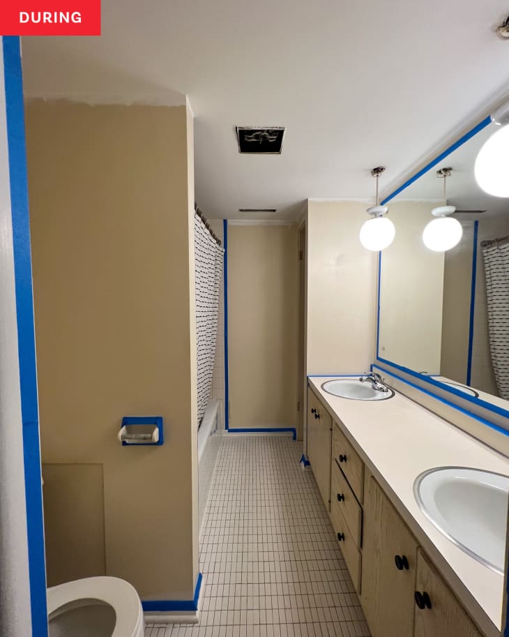 Bathroom during repainting/renovation: white and cream walls, blue painters tape around trim, mirror, etc to prepare for new paint