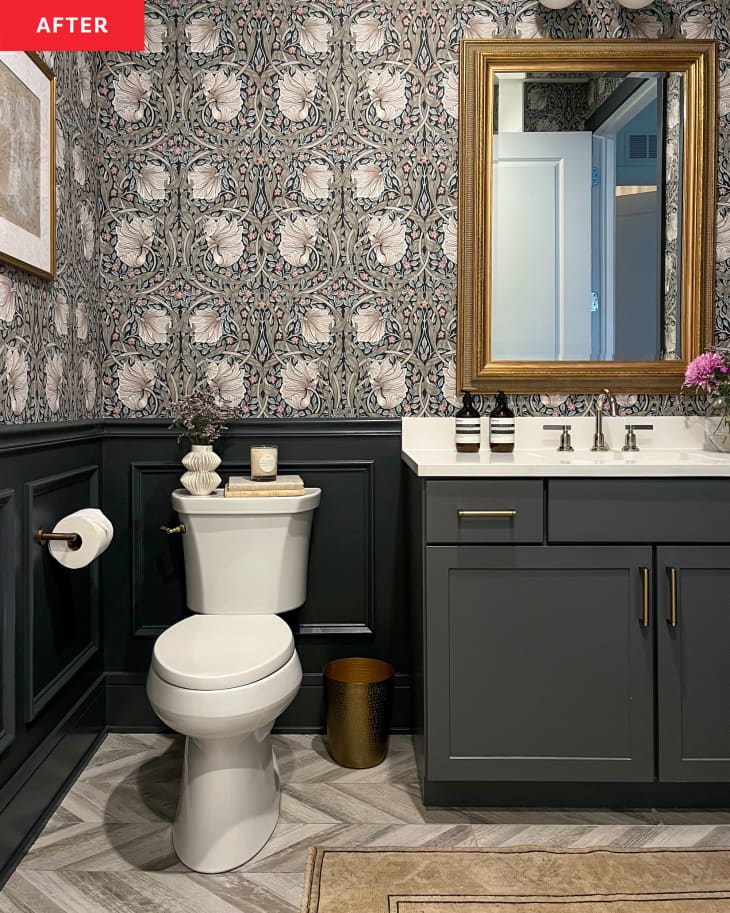 Gold mirror mounted over dark vanity with light countertops in newly renovated bathroom with dark painted wainscoting and floral wallpaper.