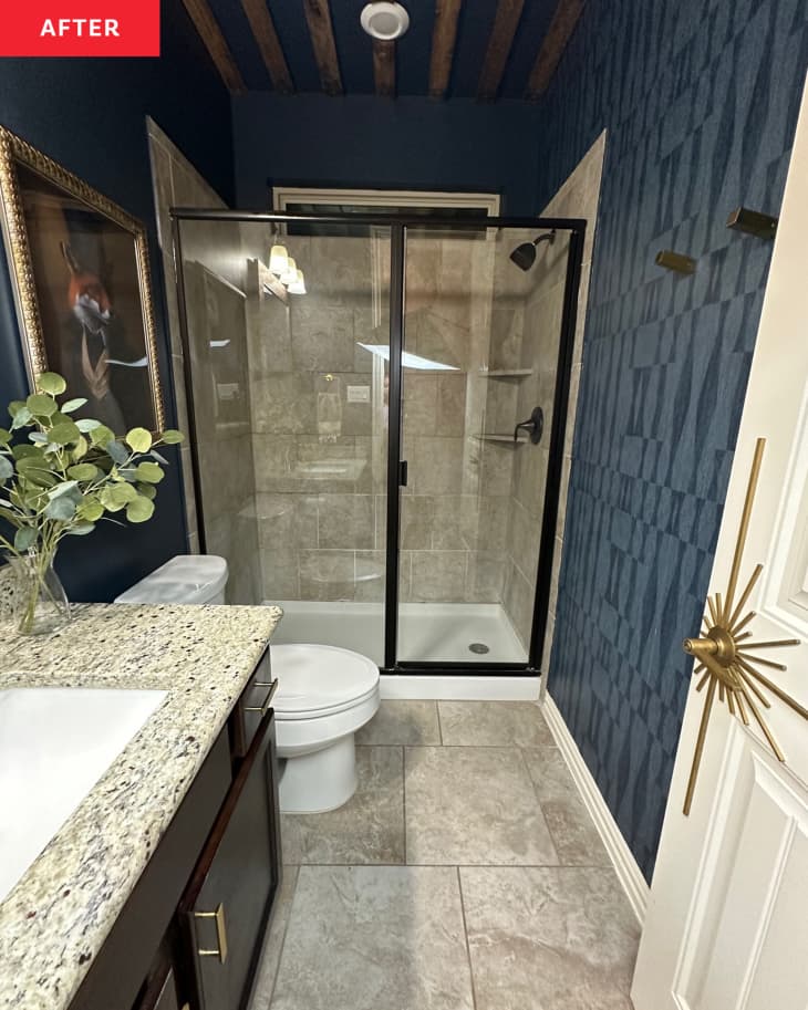 Glass shower door with black frame in blue painted bathroom with monotone graphic wallpaper and wooden planks added to ceiling.