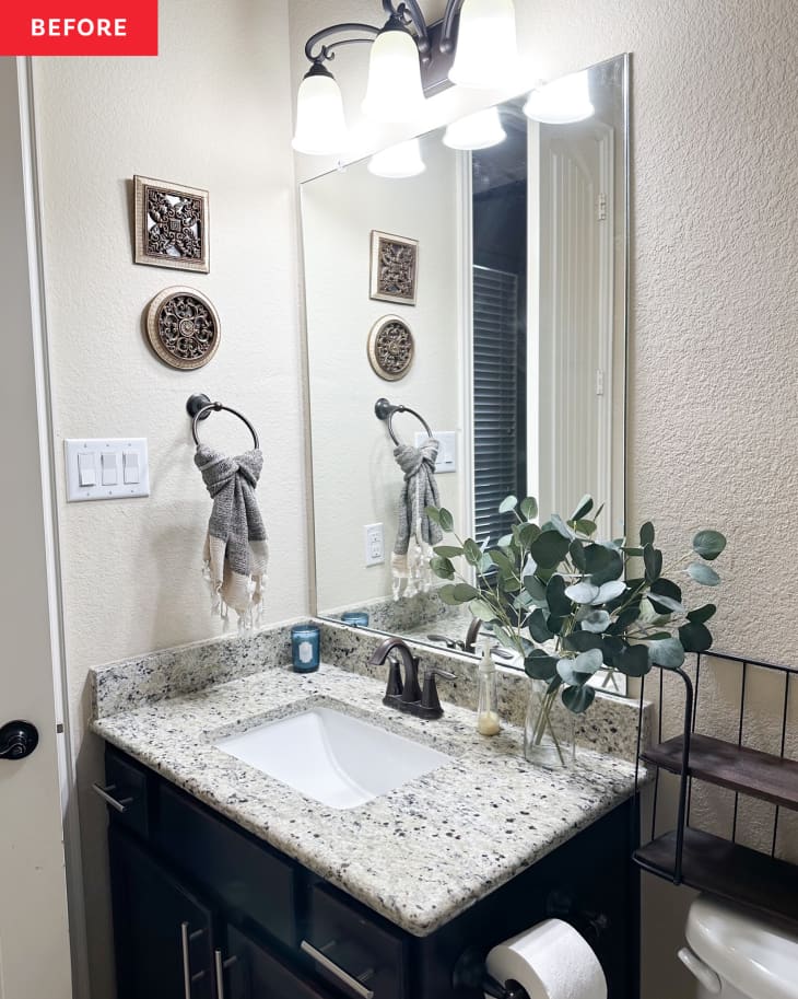 Rectangular mirror above dark wood bathroom vanity with grey and beige speckled stone surface. Small vase of eucalyptus on vanity top.