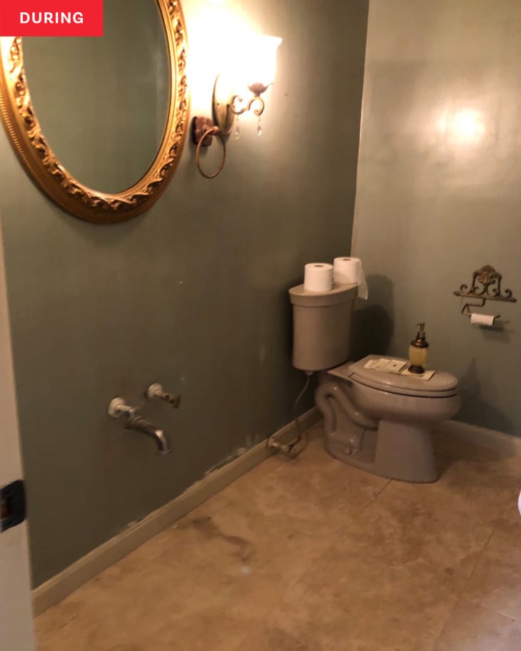 Gold round mirror and toilet remain in bathroom after vanity is removed during renovation.