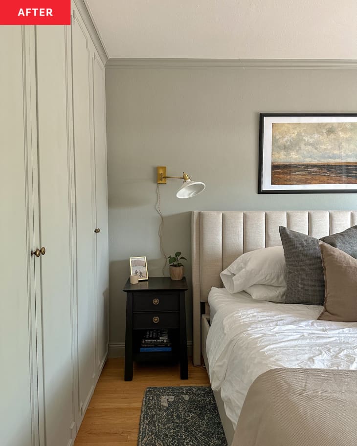 Primary bedroom after renovation: Bed with quilted gray headboard, sliding glass doors letting in light with open pale neutral curtains, pale nuted green walls, framed painting over bed, new built in floor to ceiling closet, small stool with plant on it. Bed has bedding in grays and browns, lots of pillows