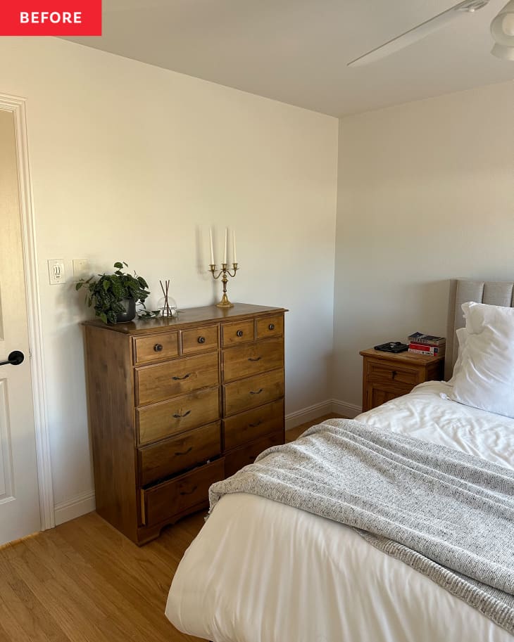 Primary bedroom before renovation: White walls with no art or decoration, wood dresser against wall with a plant, candles, bed with quilted gray headboard, sliding glass doors letting in light with open pale neutral curtains