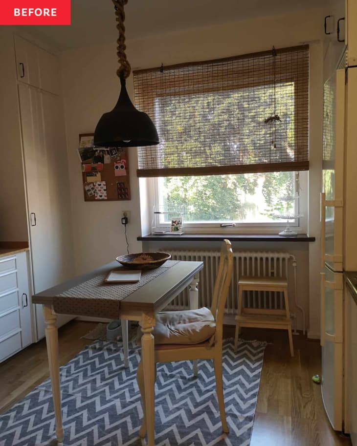 grey and white zig zag stripe rug, black hanging light fixture with chain cover, wood desk table with one wood chair, beige chair cushion, wood stool, white walls, white radiator, bamboo shade, cork board, wood floors