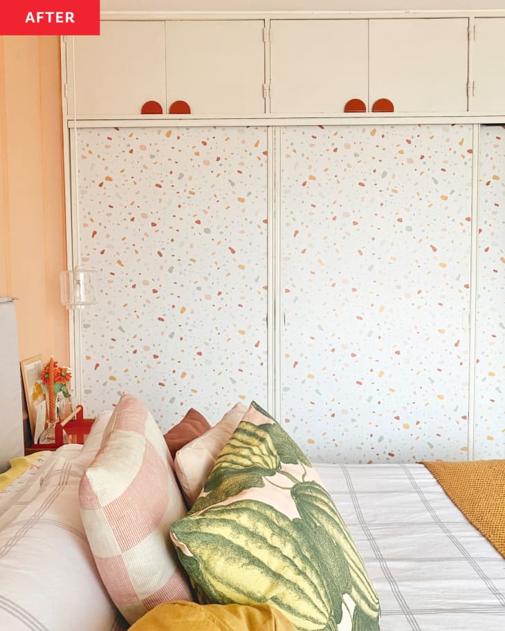 bedroom after repainting/remodel. Cantaloupe colored walls with decorative moulding, art prints of lamps, wood nightstand, white, ochre, blush, rose bedding with watermelon pattern pillow, cloest doors with terrazo pattern in white, gray, blush tones
