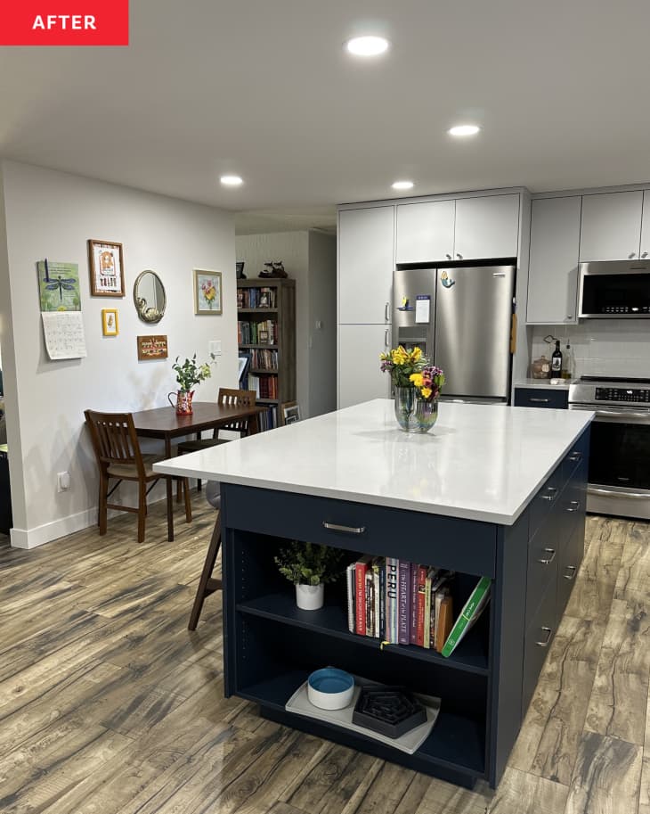 large island with built in book shelves, white counter top, floor to ceiling built in cabinets around fridge, wood floors, small table for two, gallery wall with a few pieces of art