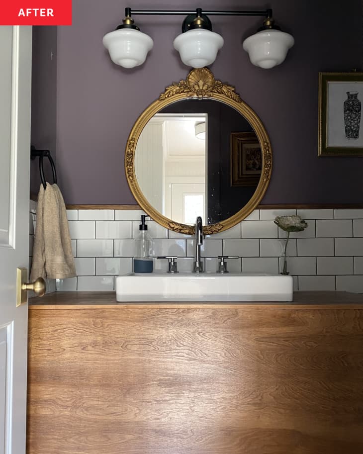 large wood vanity, raised white sink bowl, purple walls, 3 light scones fixture above mirror, round gold mirror, white subway tile backsplash