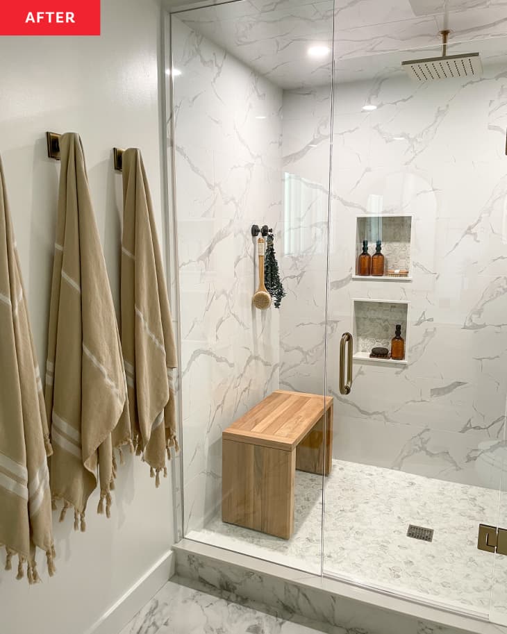 Beige towels hung near a white stone shower with glass doors and a wooden bench.