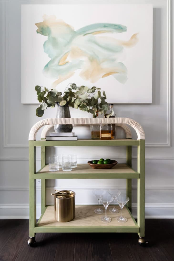 Light green bar cart with rope-wrapped railings, filled with glasses, bar ware, and bottles.