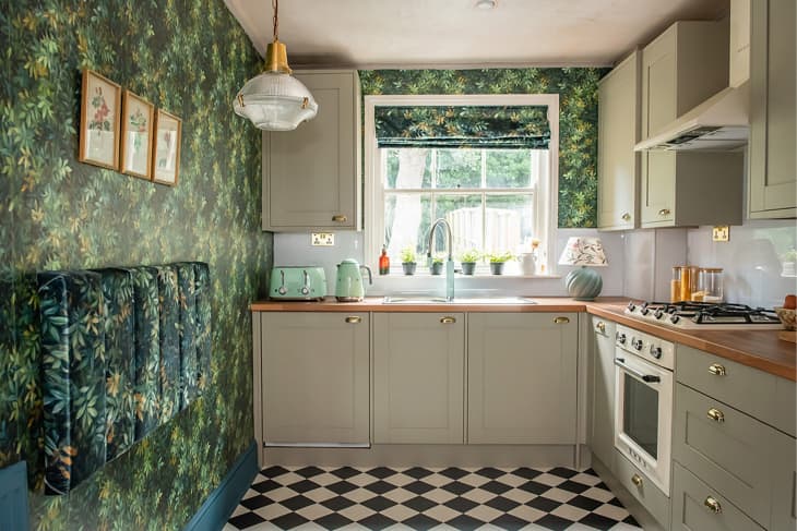 Kitchen with floral wallpaper, wooden countertop, and light green cabinets with golden hardware.
