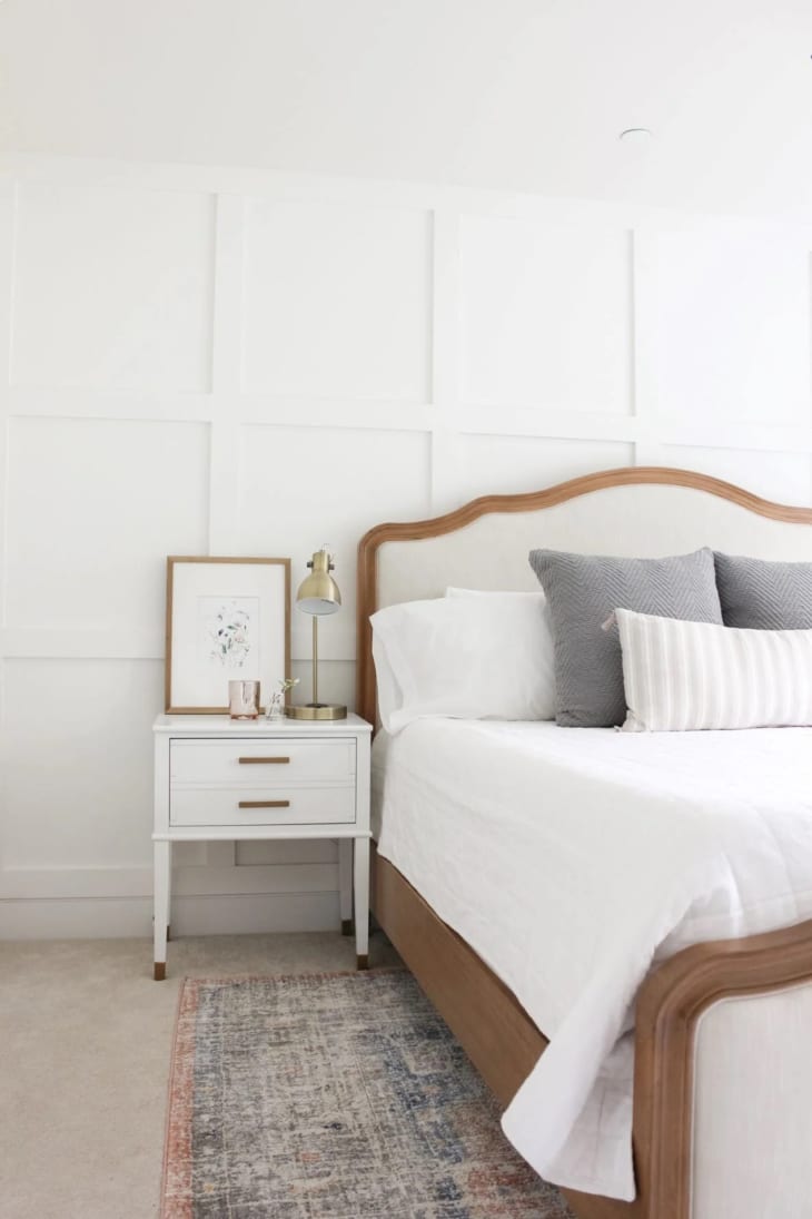 white wainscoting on wall, white fabric bed with wood trim, small white nightstand