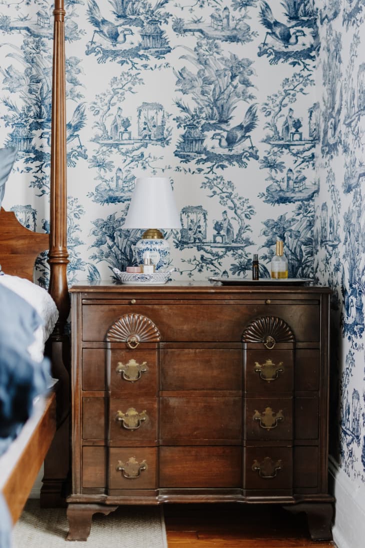 Blue toile wallpaper, dark wood dresser with small blue and white ceramic lamp on top, wood four poster bed