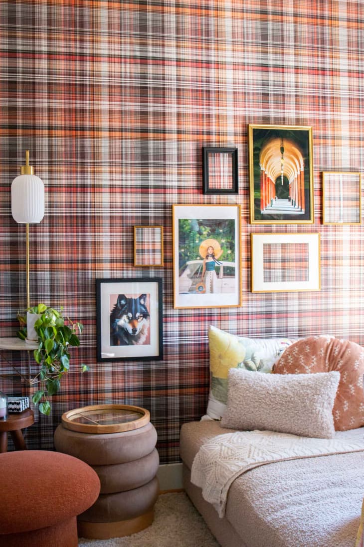 Framed pieces of art on a plaid wallpapered wall next to a fuzzy couch with decorative pillows.