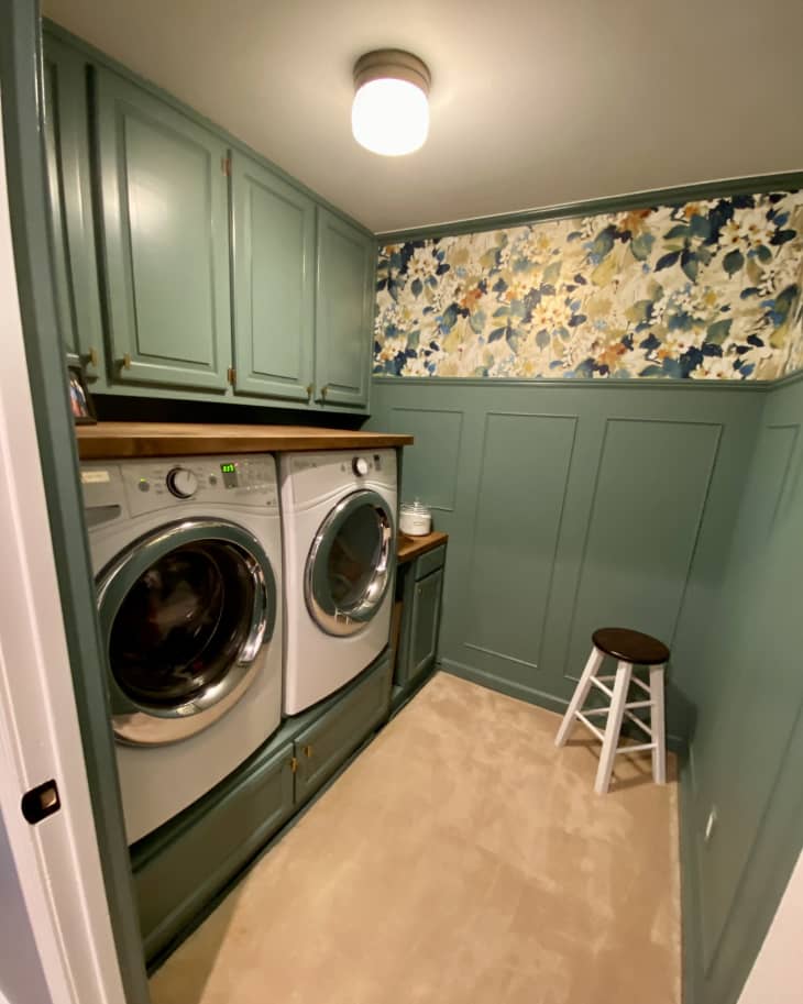 Green wainscoting, bold floral wallpaper on upper half of wall, countertop built on top of washer and dryer with built in cabinets above, small wood and white stool in corner
