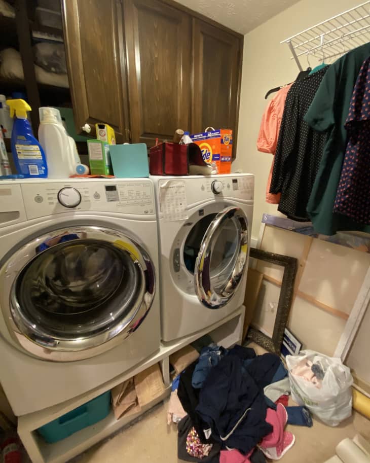 Wooden cabinets with washer and dryer underneath, messy top with detergent and cleaning supplies everywhere, clothes hanging on rack, pile of clothes on floor