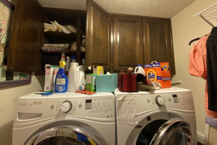 Wooden cupboards with washer and dryer underneath, messy top with detergent and cleaning supplies throughout