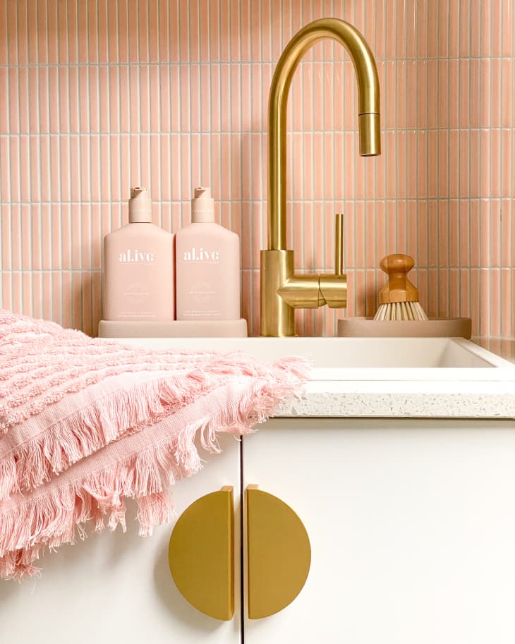 Laundry room with pink tiles, white cabinets with gold round pulls, and other gold details after renovation. Detail of sink area