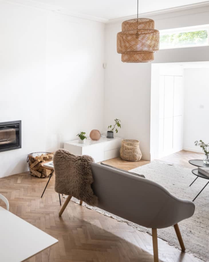 Basket of logs next to fireplace without mantel in light living room with corner storage, light seating, and rattan lamp.