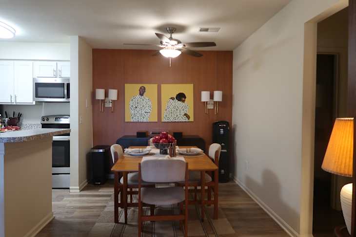 Dining area featuring a wooden table set for four, two wall art pieces, and a modern kitchen in the background.
