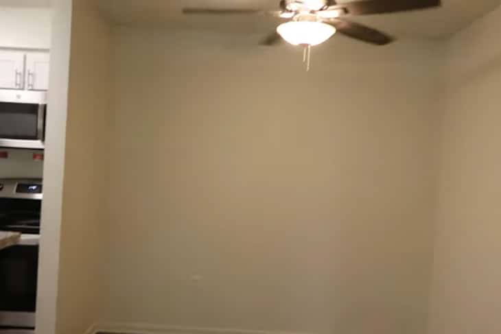 Small room with a ceiling fan, light fixture, and a view of a kitchen area with stainless steel appliances.