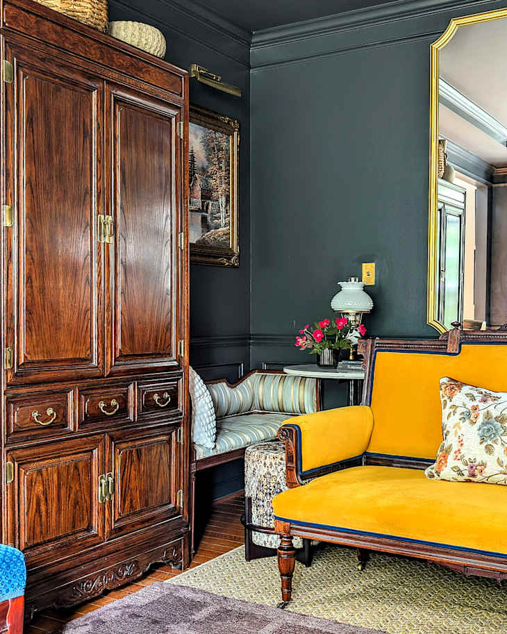 Cozy living room corner featuring a yellow upholstered sofa, wooden armoire, striped bench, and floral decor.