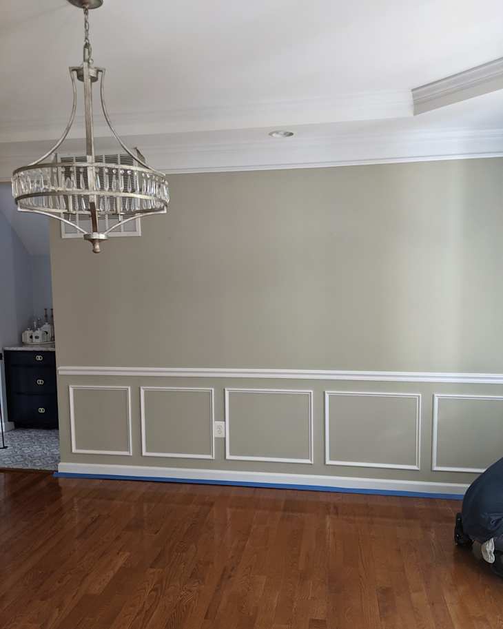 Chandelier hanging from the ceiling above a light green wall with white wainscoting and hardwood floor.