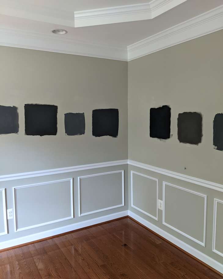 Wall with several black paint swatches on a light gray background, featuring white wainscoting and hardwood flooring.