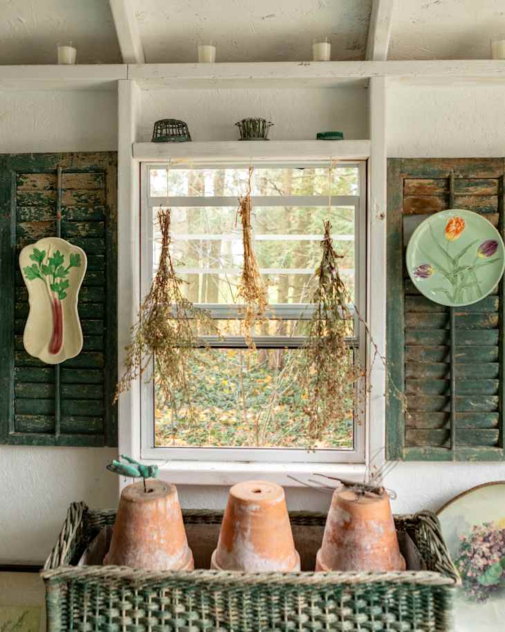 Woven basket with terracotta pots, dried herbs hanging by a window, and decorative plates on green shutters.