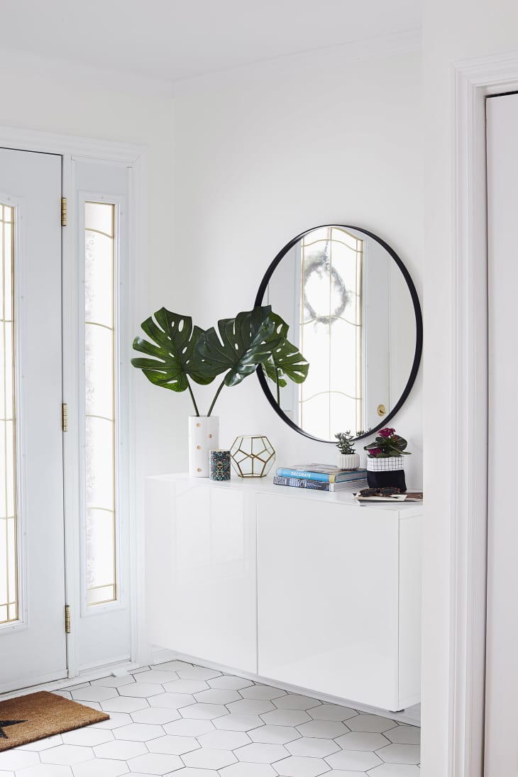 A bright white entryway with a storage cabinet and a round mirror on the wall with Monstera plant leaves in a vase as decor