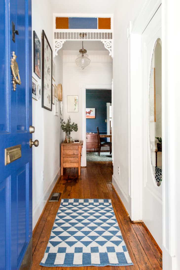 A bright blue door opening up into a tall, narrow entryway with white walls, a wood floor, and a geometric-patterned rug