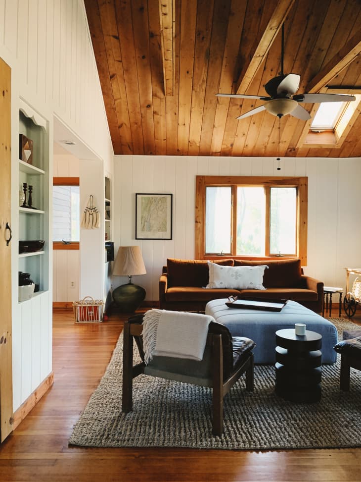 Cabin living room with tall wood ceilings, brown sofa, blue ottoman, and warm rug.