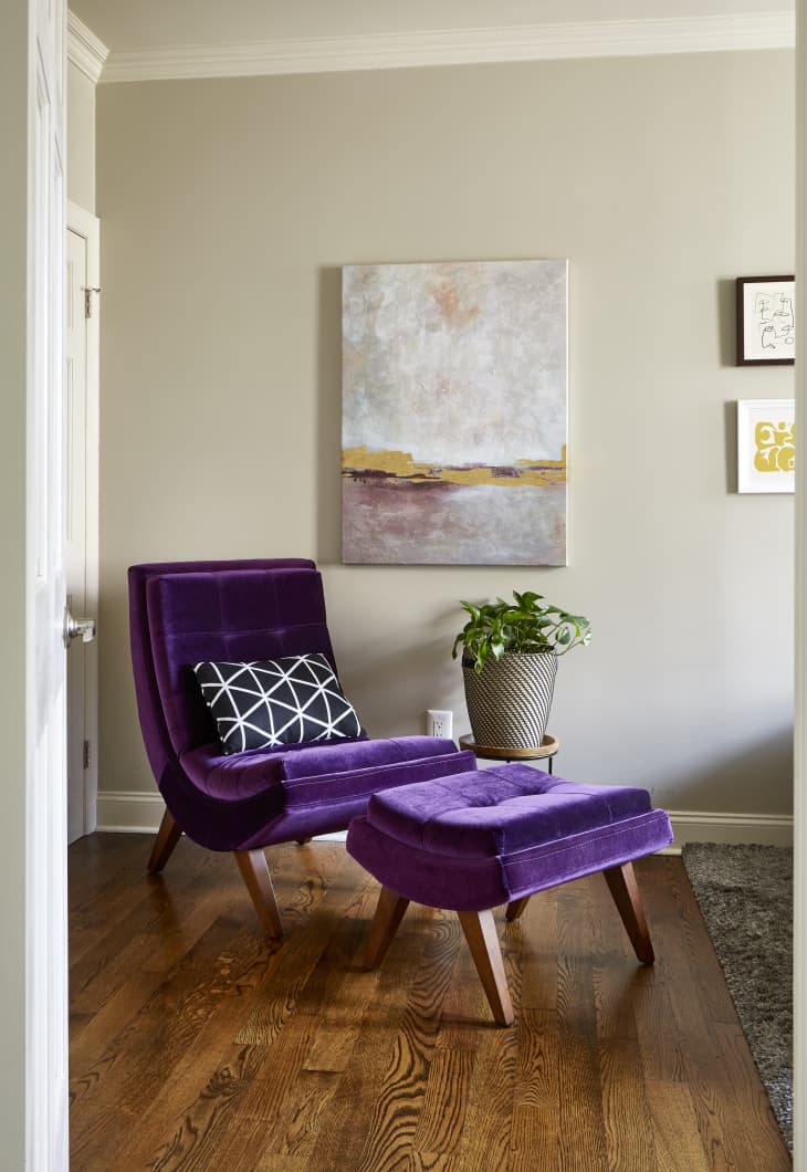 Modern purple velvet armless lounge chair and ottoman in a corner, with a plant and abstract art