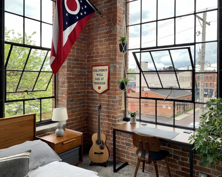 Guitar, flag, desk, and nightstand with lamp in bedroom with large windows