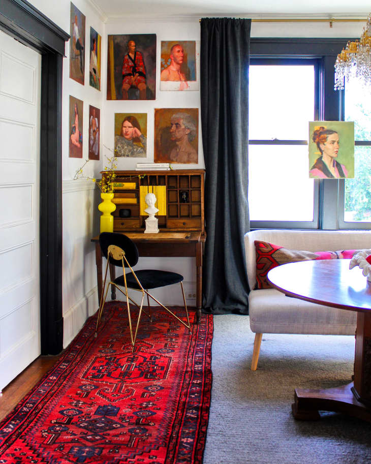 Desk in corner of dining room with painted portraits hanging on wall, black curtains, and round wooden table