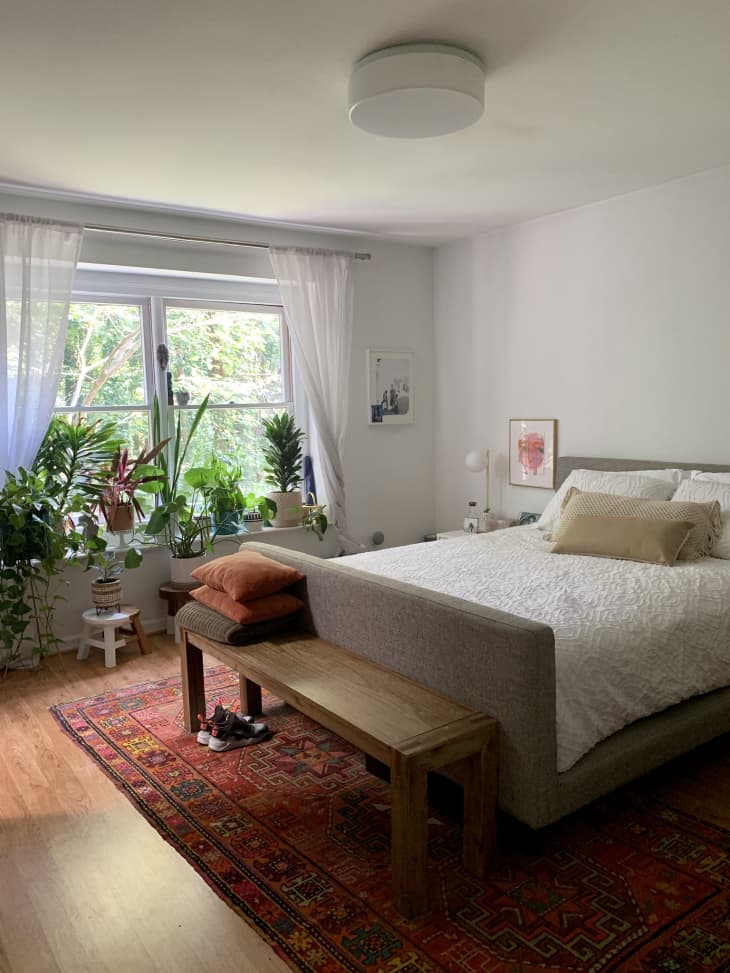 Bedroom with large window, lots of plants, white bedding and walls, and red-orange rug