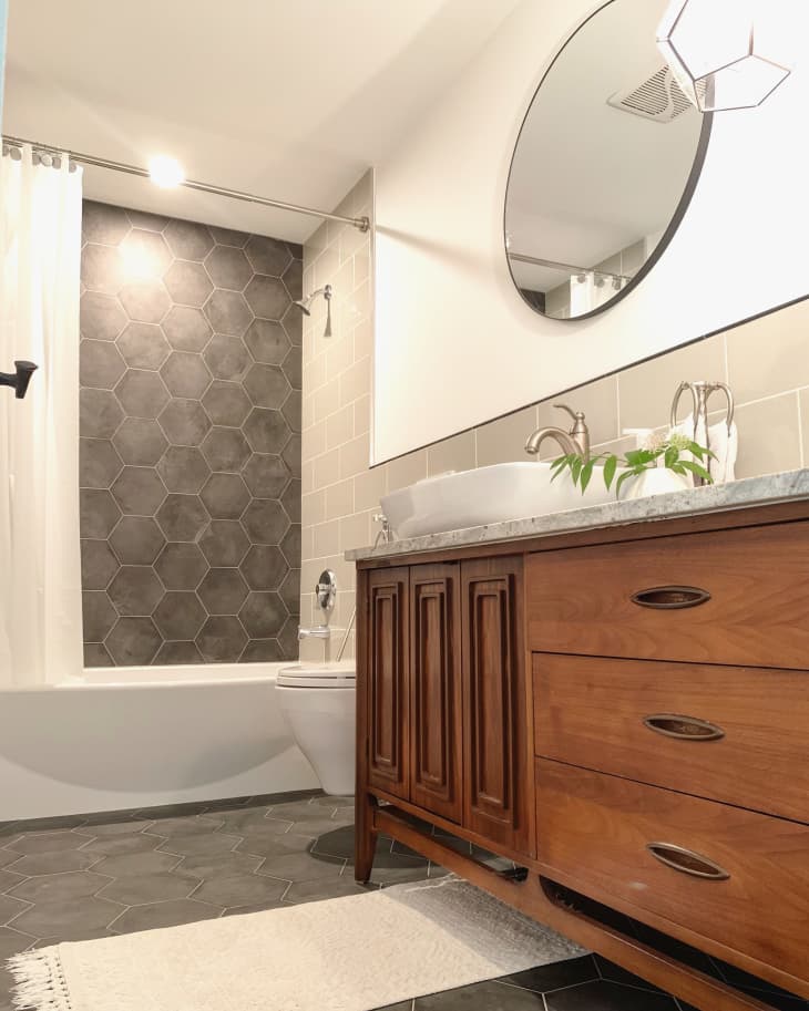 Bathroom with gray hexagonal tile in shower and circular mirror above mid-century vanity
