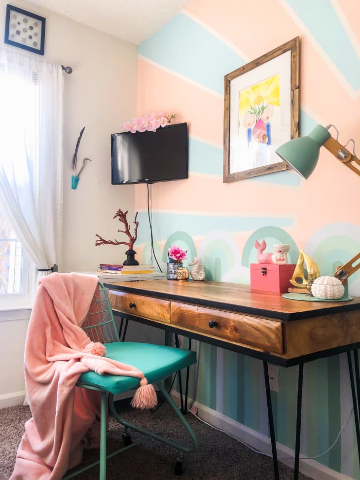 Desk with blue-green chair, blue-green lamp, and pink and blue mural on wall behind it