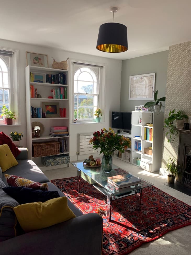 Sunlit living room with gray sofa, red rug, bookshelf between two windows, and black drum-shaped pendant light