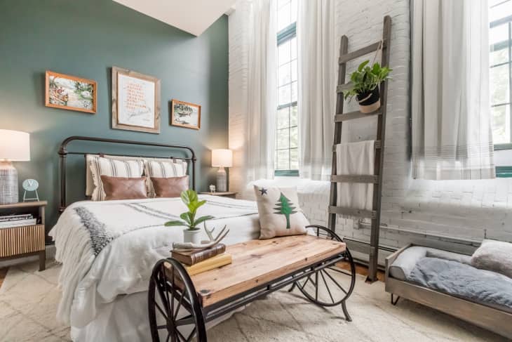 Bedroom with green walls, exposed brick painted white, and antique bench with wheels at foot of bed