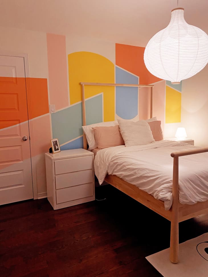 Colorful bedroom with wooden IKEA bed, geometric colorful mural painted on the bed wall and across the door, and a large white hanging paper pendant