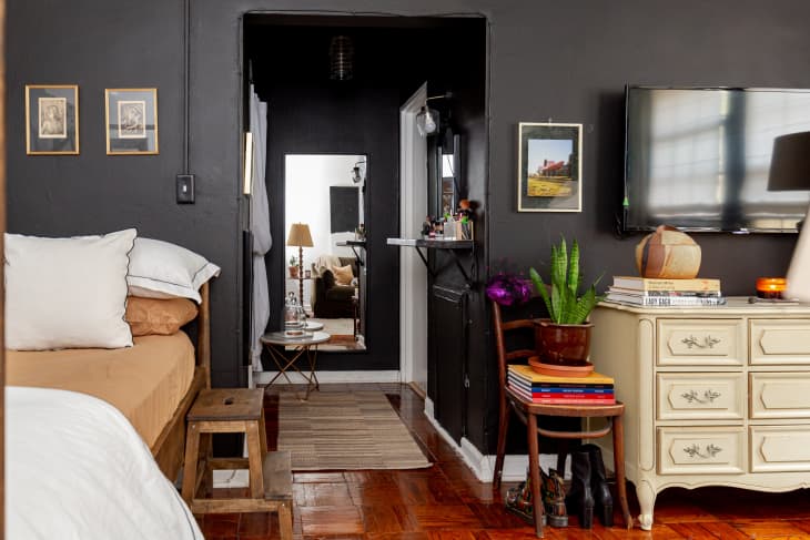 Bedroom with a beige bed, wooden stool, dresser, potted plant, and wall art, leading to a hallway with a mirror.