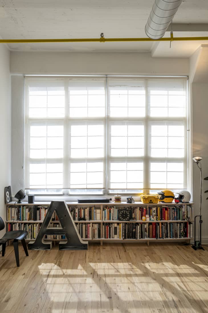 A large industrial wall of windows in a modern, minimal loft apartment, with a long low shelf of books and a large letter 'A' sculpture.