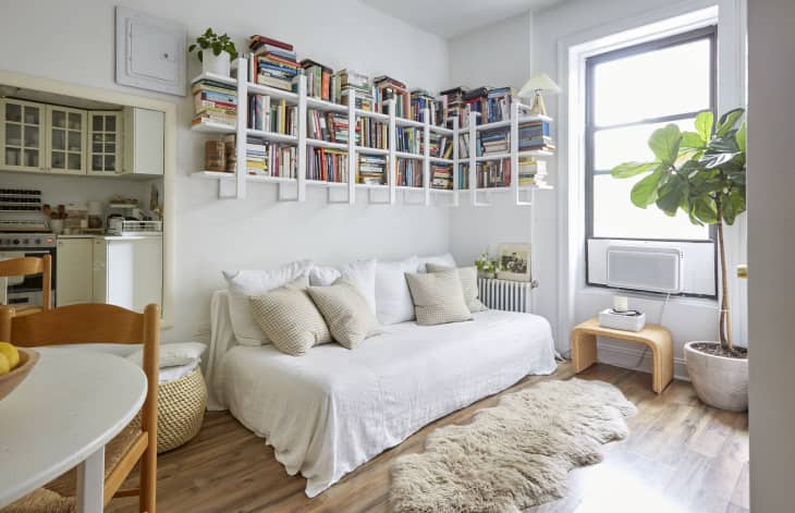 Cozy living room with white sofa, wall-mounted bookshelves, large window, and a potted plant on wooden floor.