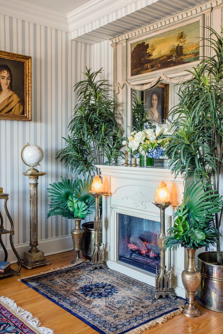 Elegant living room with striped wallpaper, ornate fireplace, lush plants, vintage portraits, and decorative globe.