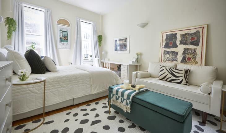 Bright bedroom with a white bed, teal ottoman, and cream sofa. Art of roaring cats on the wall, and hanging plants by windows.
