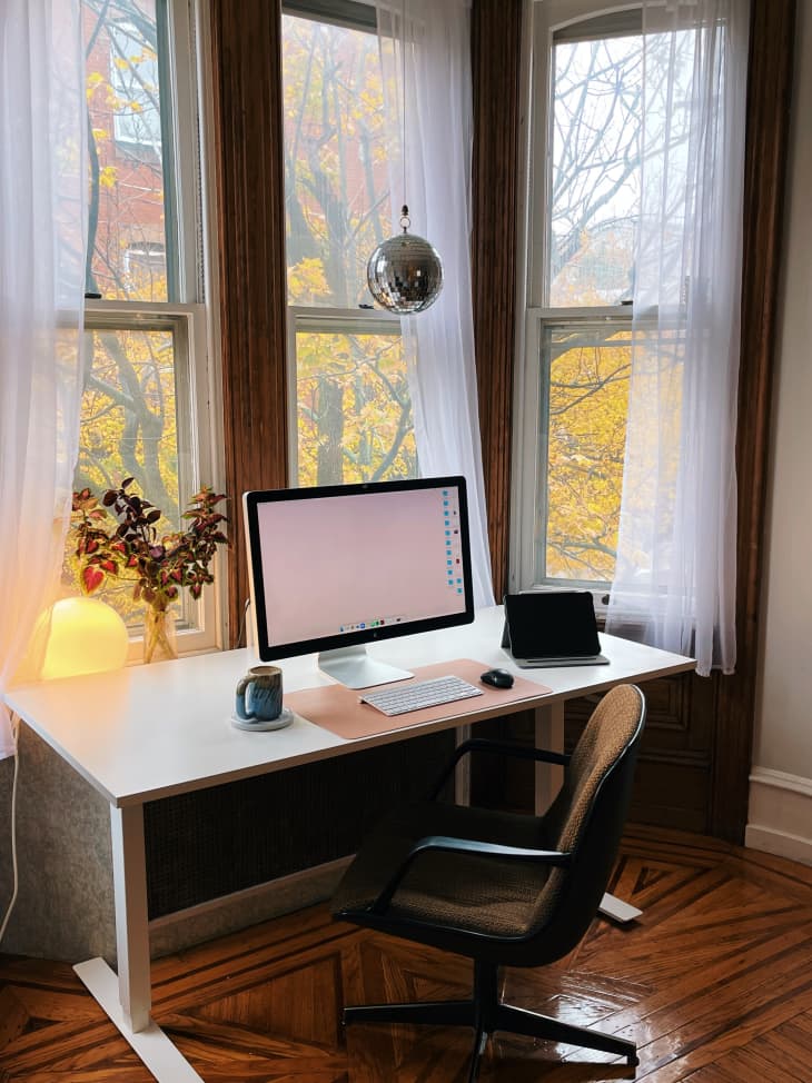 A white desk next to three tall windows with white curtains