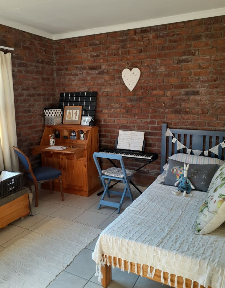 The corner of a brick bedroom with a desk, piano, and bed