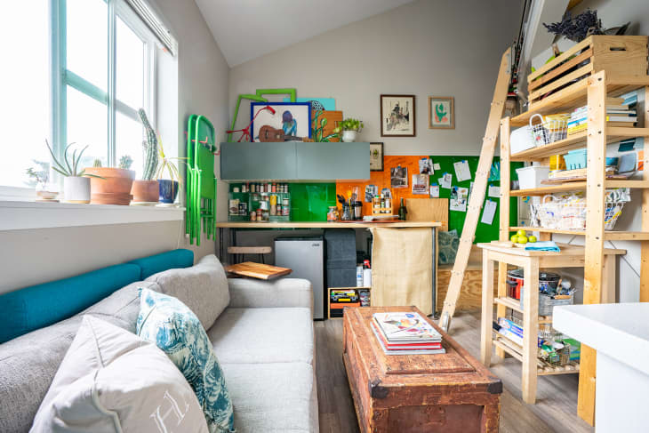 Cozy studio with a gray sofa, wooden shelves, colorful wall art, and a small kitchenette with green and orange accents.