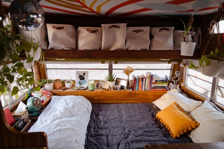 一辆上世纪70年代露营车的卧室角落，床上方有架子和篮子
