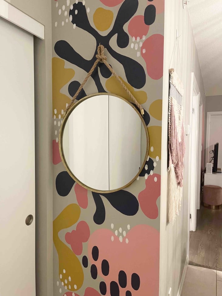 A wall by a closet painted with blue, pink, and yellow leaves and spots with a round mirror hanging over it